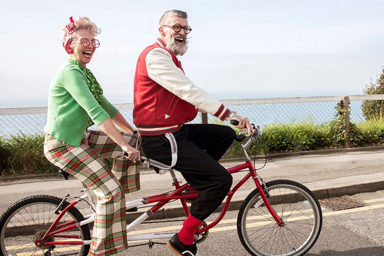 Zwei ältere Menschen sitzen lächelnd auf einem Tandem-Fahrrad.