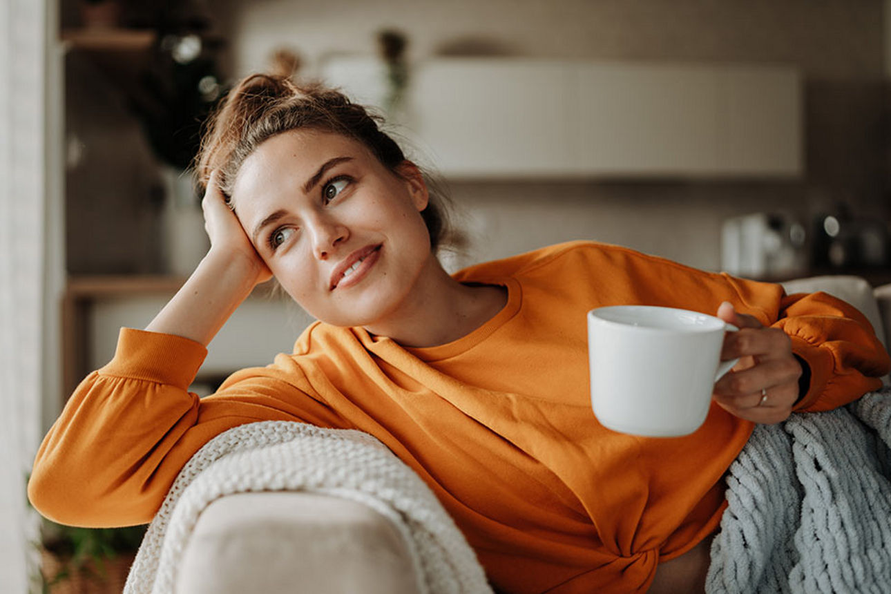 Person lehnt mit dem Kopf auf der Hand auf einem Sofa und hält eine weiße Tasse in der anderen Hand