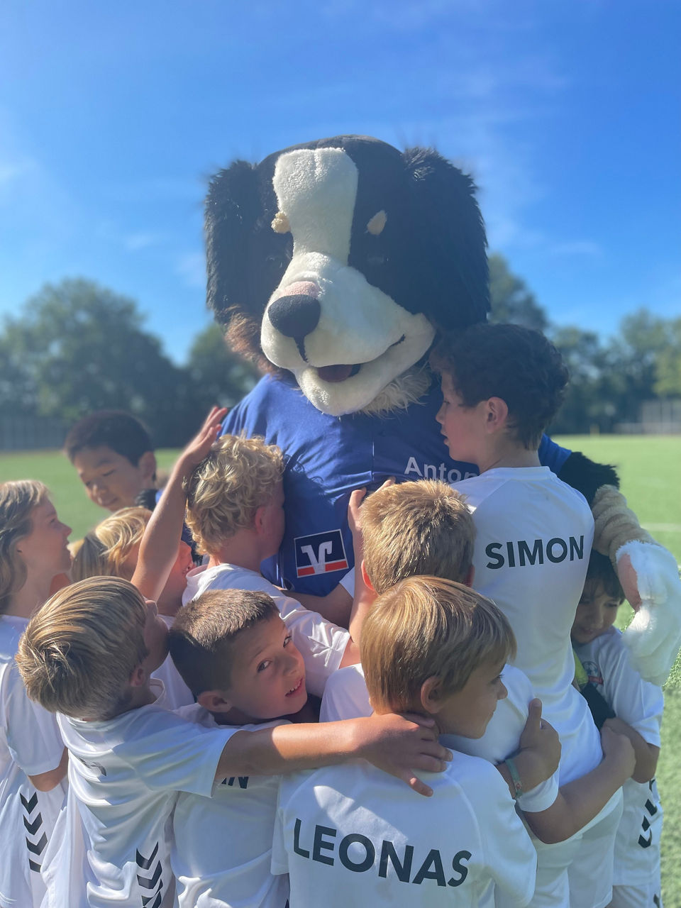 Unser Maskottchen (Bankhund Anton) wird von mehreren Kindern in Sporttrikots umarmt.