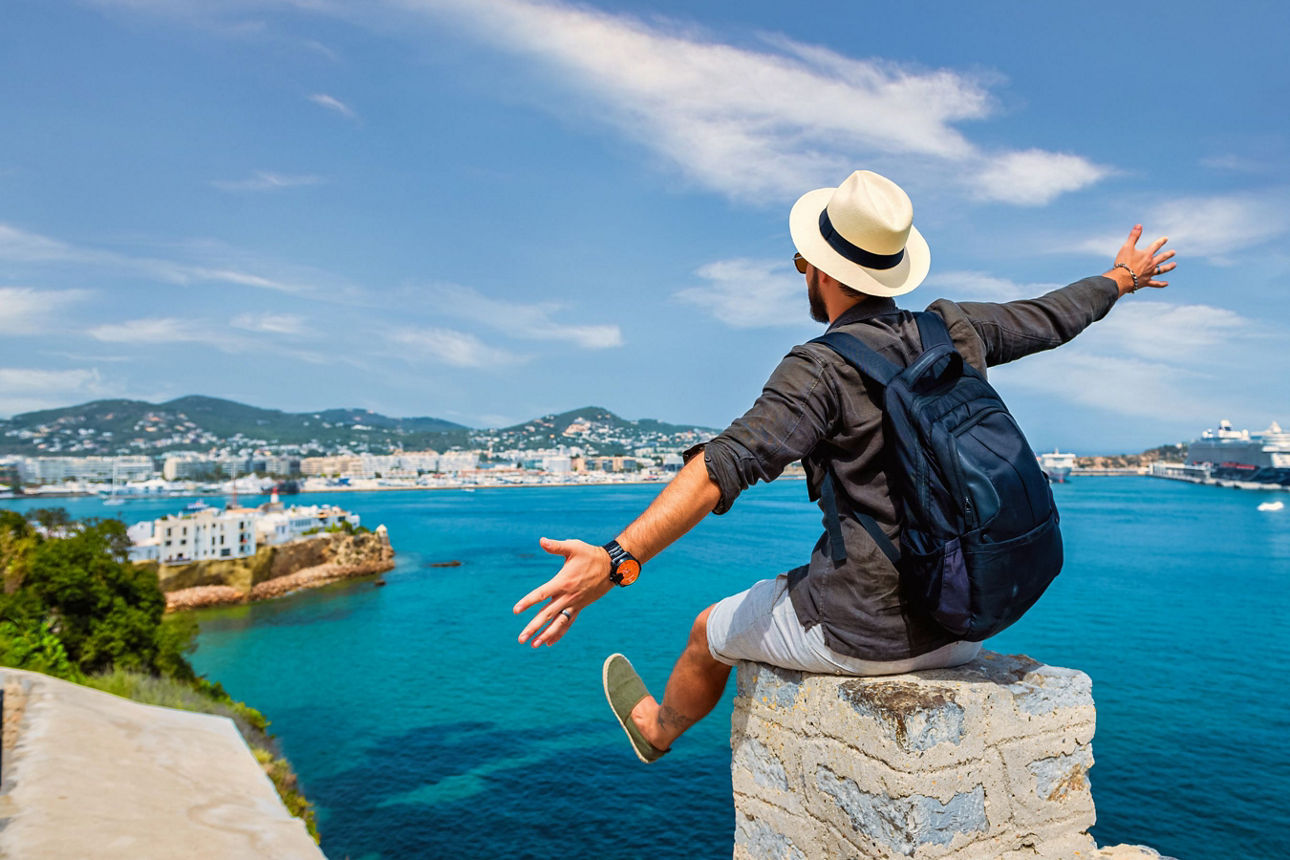 Ein Mann mit Hut und ausgestreckten Armen sitzt auf einer Mauer mit Sicht auf das blaue Meer