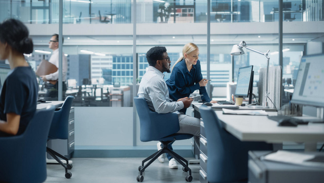 Frau schaut am Schreibtisch sitzenden Mann auf den Monitor