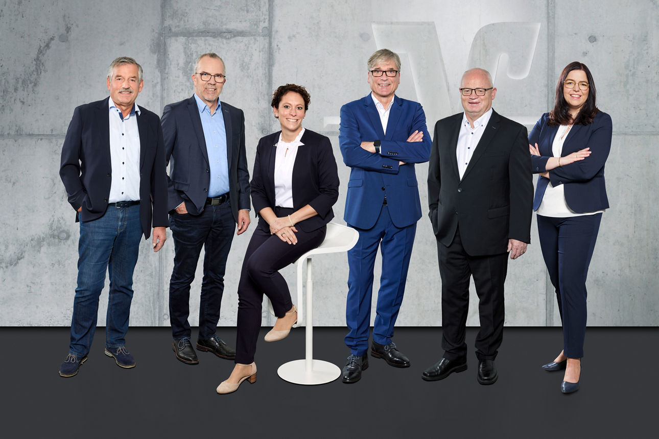 Es sind vier Männer und zwei Frauen in Business-Kleidung zu sehen. Sie lächeln in die Kamera.
