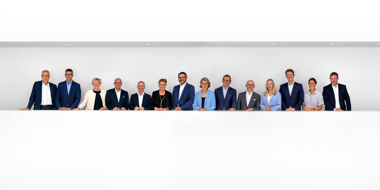 Vierzehn Personen in Businesskleidung stehen nebeneinander vor weißem Hintergrund.