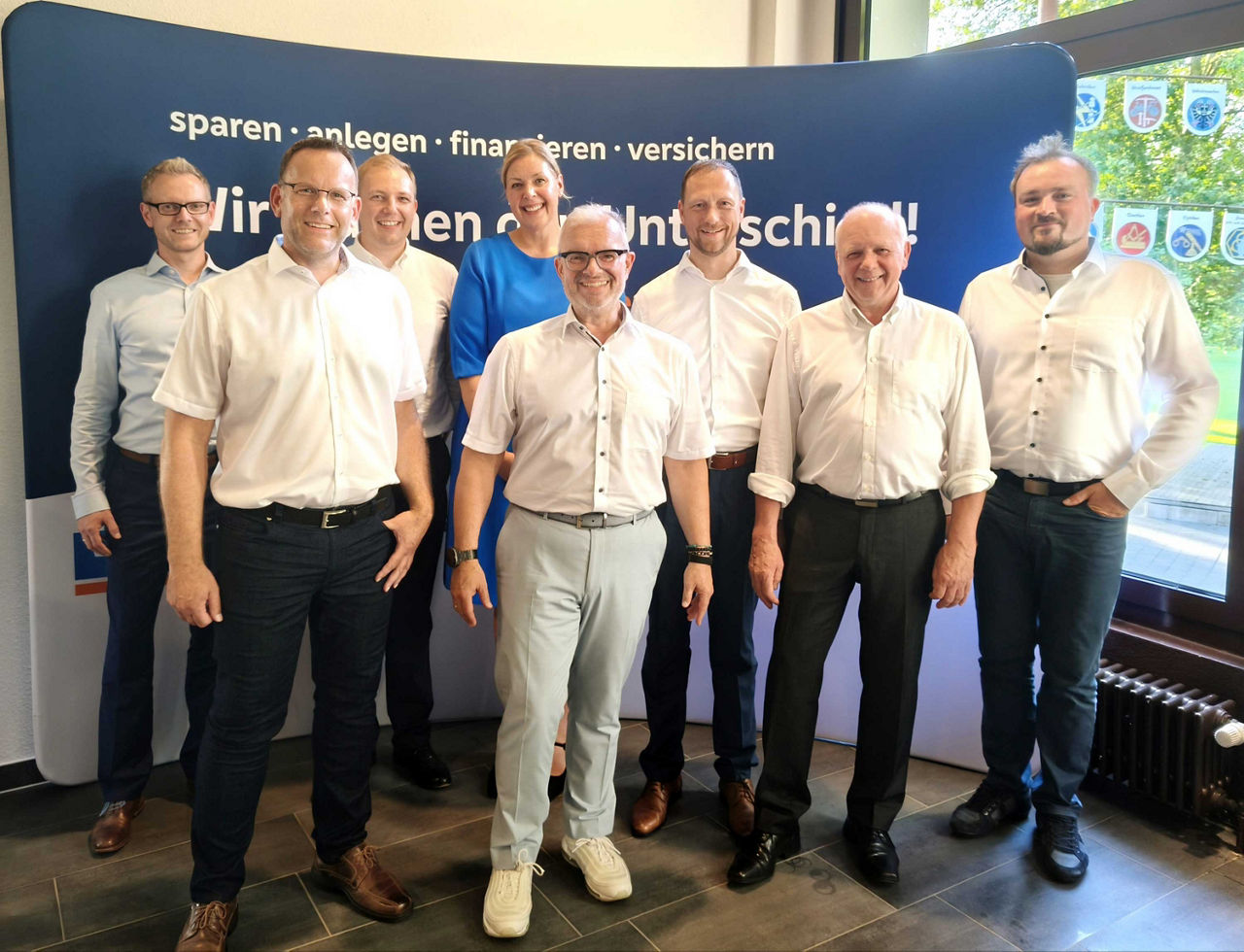 Eine Gruppe von acht Personen steht vor einer blauen Firmenwand mit der Aufschrift „sparen · anlegen · finanzieren · versiche