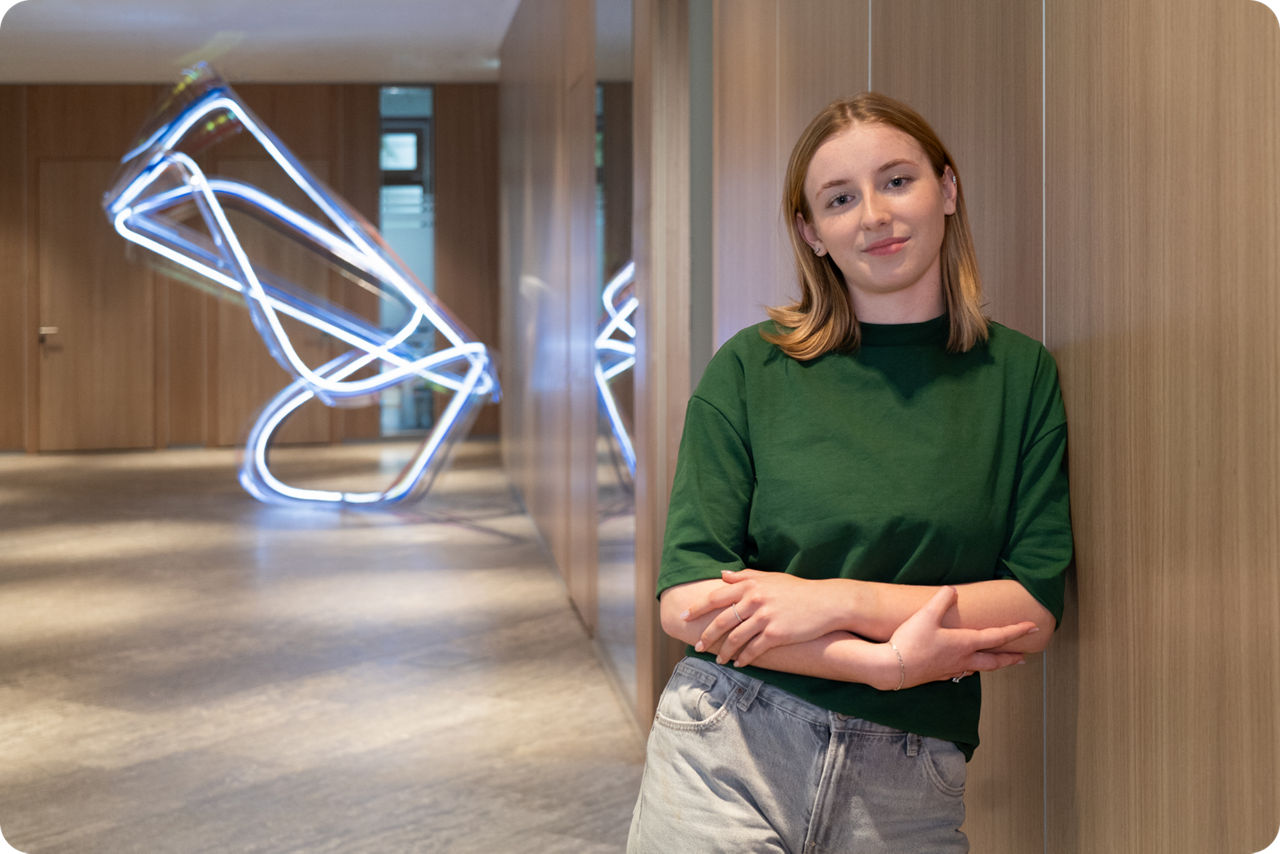 Eine junge Frau lehnt an einer Wand und lächelt in die Kamera, im Hintergrung ist eine Lichtinstallation