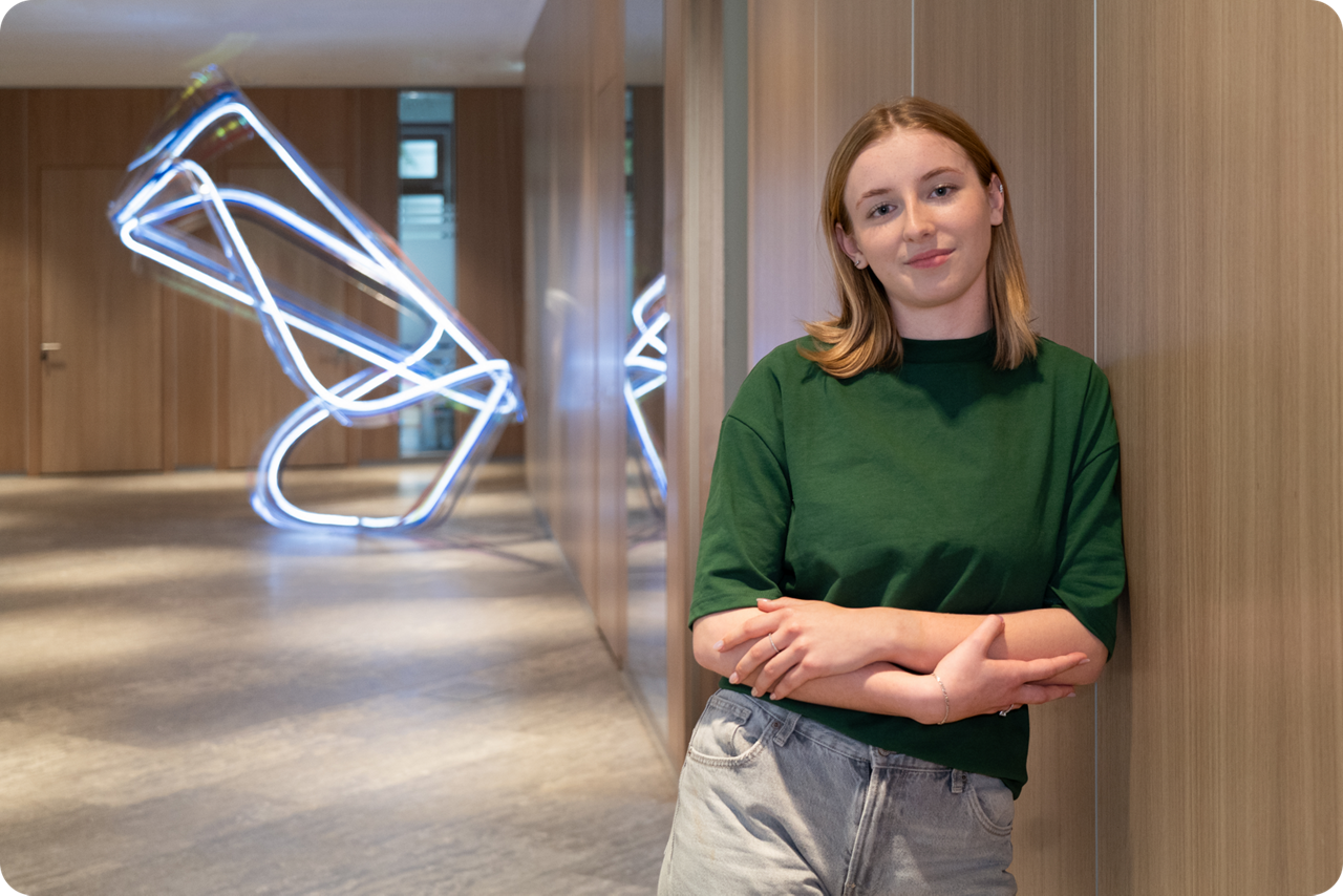 Eine junge Frau lehnt an einer Wand und lächelt in die Kamera, im Hintergrung ist eine Lichtinstallation