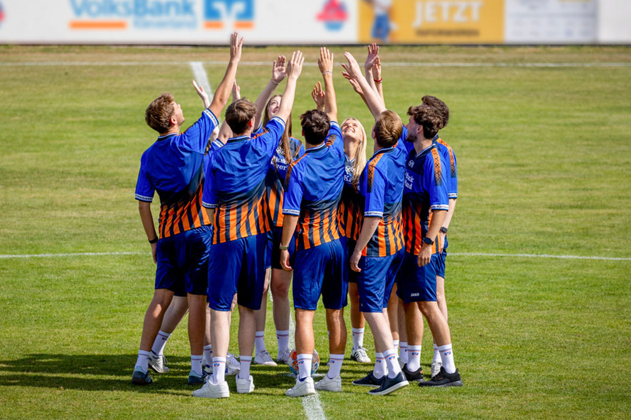 Gruppe von jungen Menschen in blauen Sporttrikots mit orangen Streifen steht im Kreis und hebt die Arme nach oben