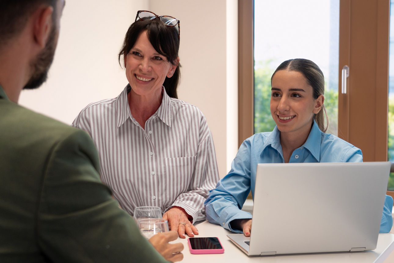 Zwei Frauen sitzen vor einem Laptop und schauen lächelnd den Gesprächspartner an