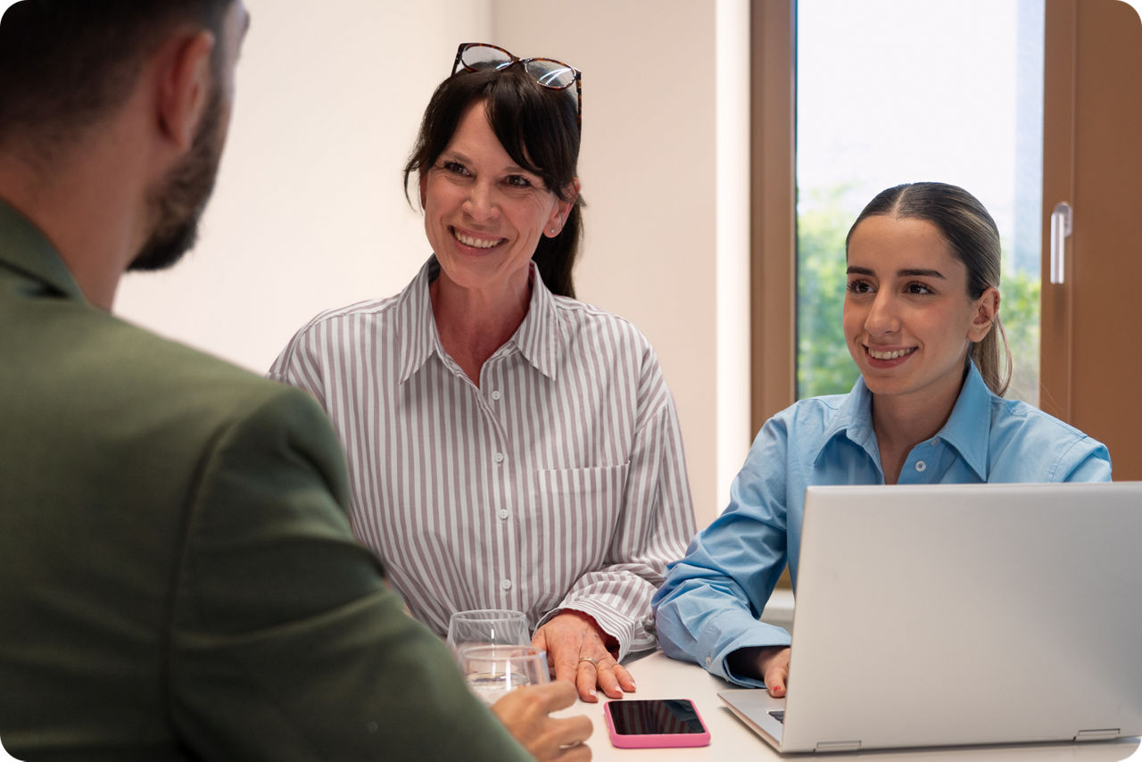 Zwei Frauen sitzen vor einem Laptop und schauen lächelnd den Gesprächspartner an