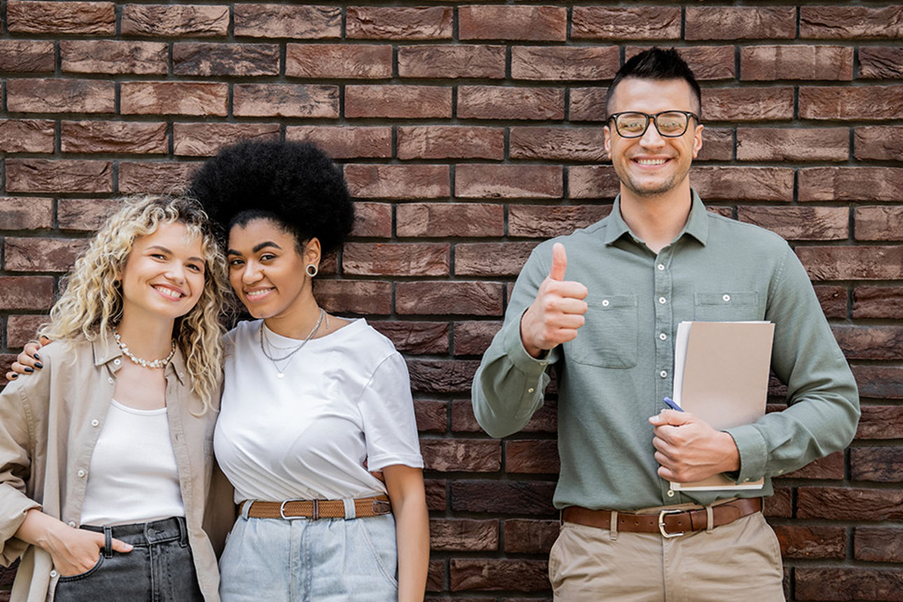 Drei junge Menschen vor einer Backsteinwand, die in die Kamera lächeln 
