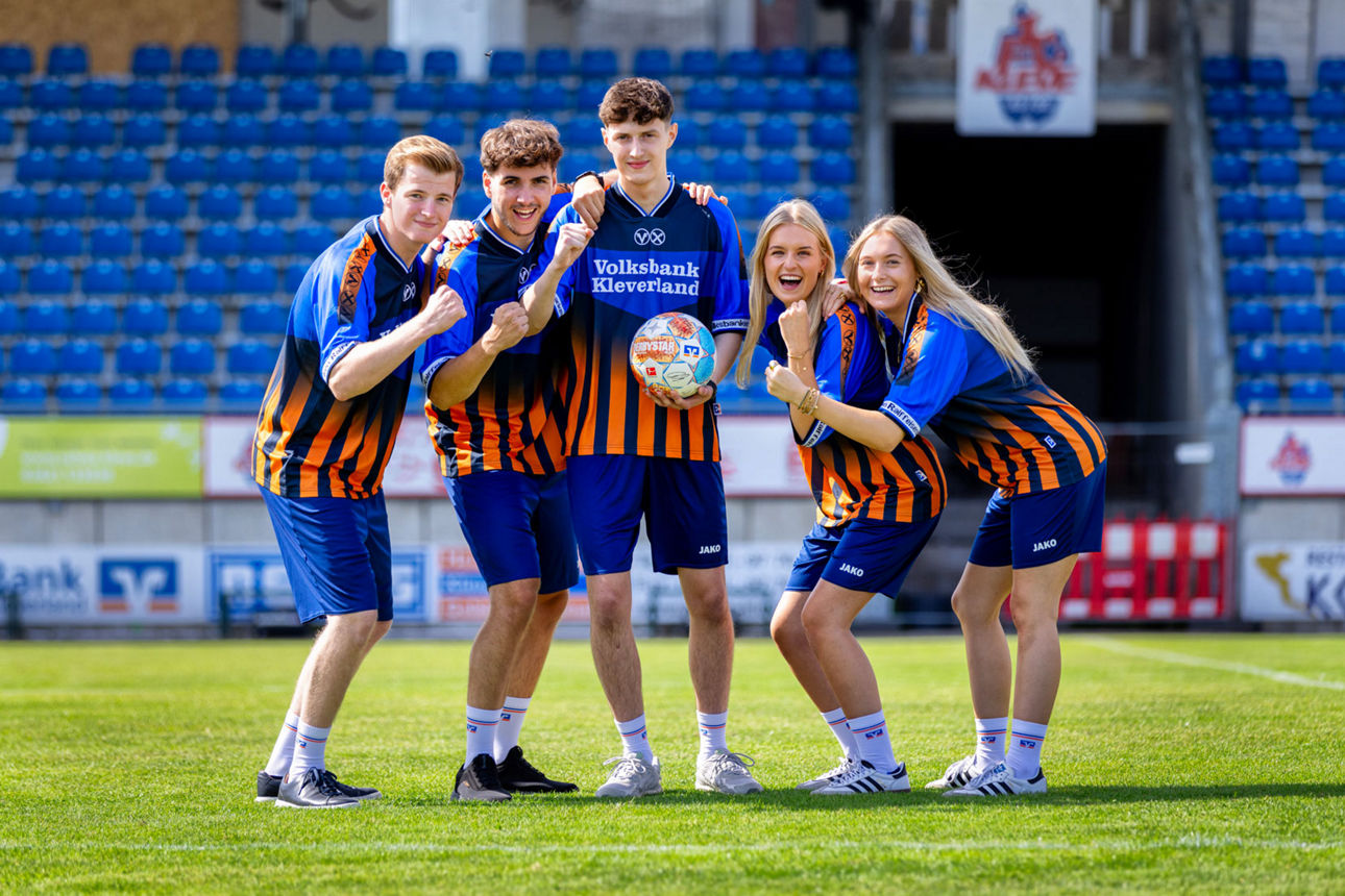 Fünf Personen in blau-orange gestreiften Trikots posieren mit einem Fusball in einem Stadion mit blauen Sitzreihen