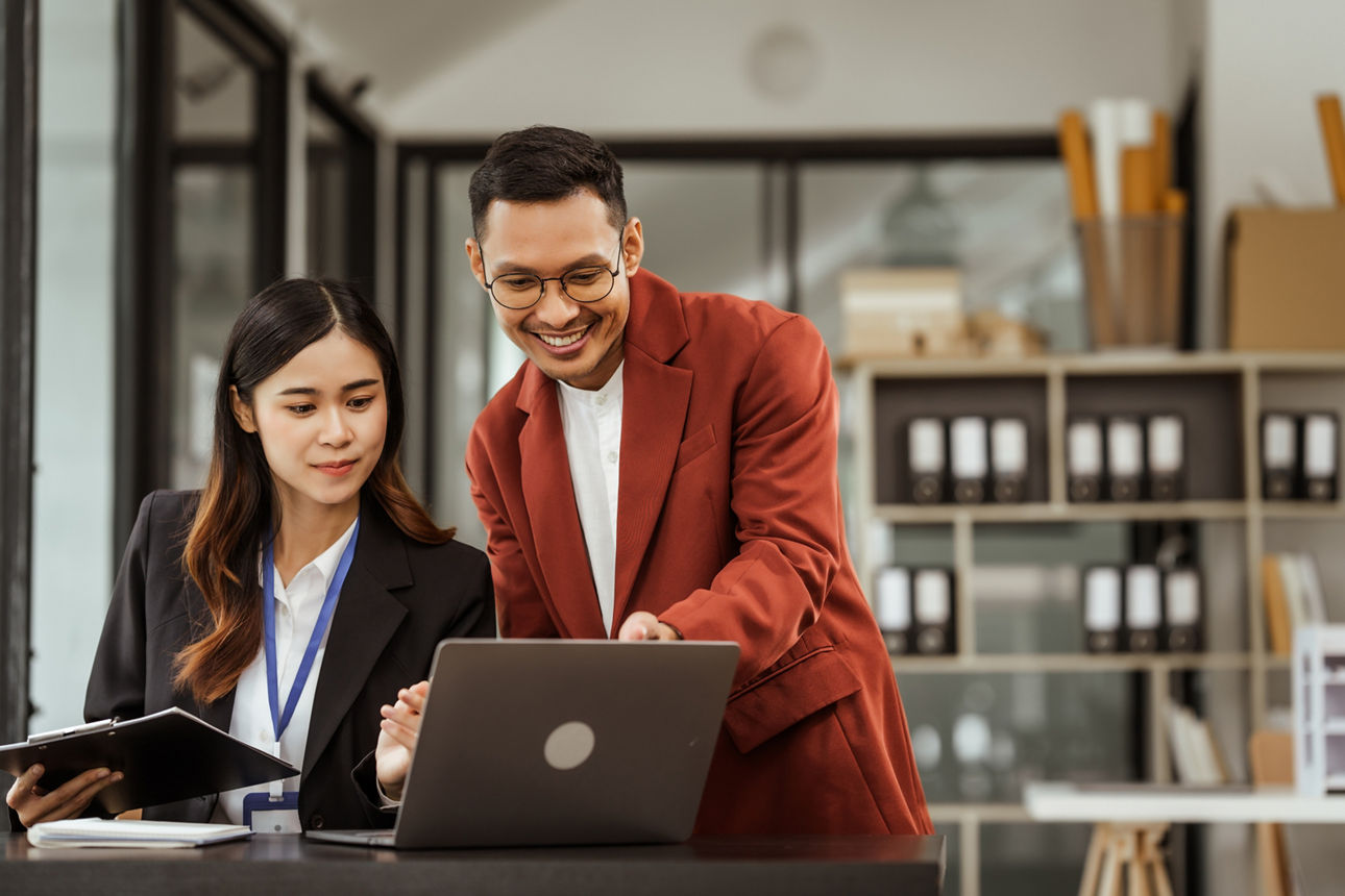 Junge Frau sitzend und junger mann stehend am Schreibtisch und schauen auf den Laptop
