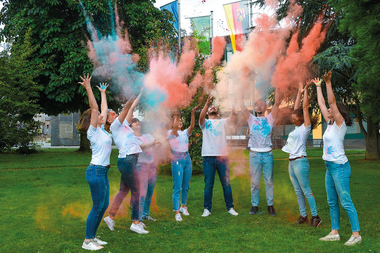 Junge Mitarbeitende der Raiffeisenbank stehen in einem Park und werfen blau-orangefarbenes Holi-Pulver hoch