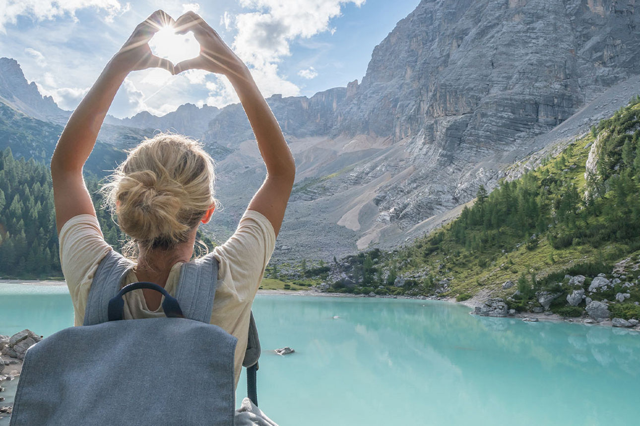 Frau blickt auf eine Berglandschaft mit See und formt mit ihren Händen ein Herz.