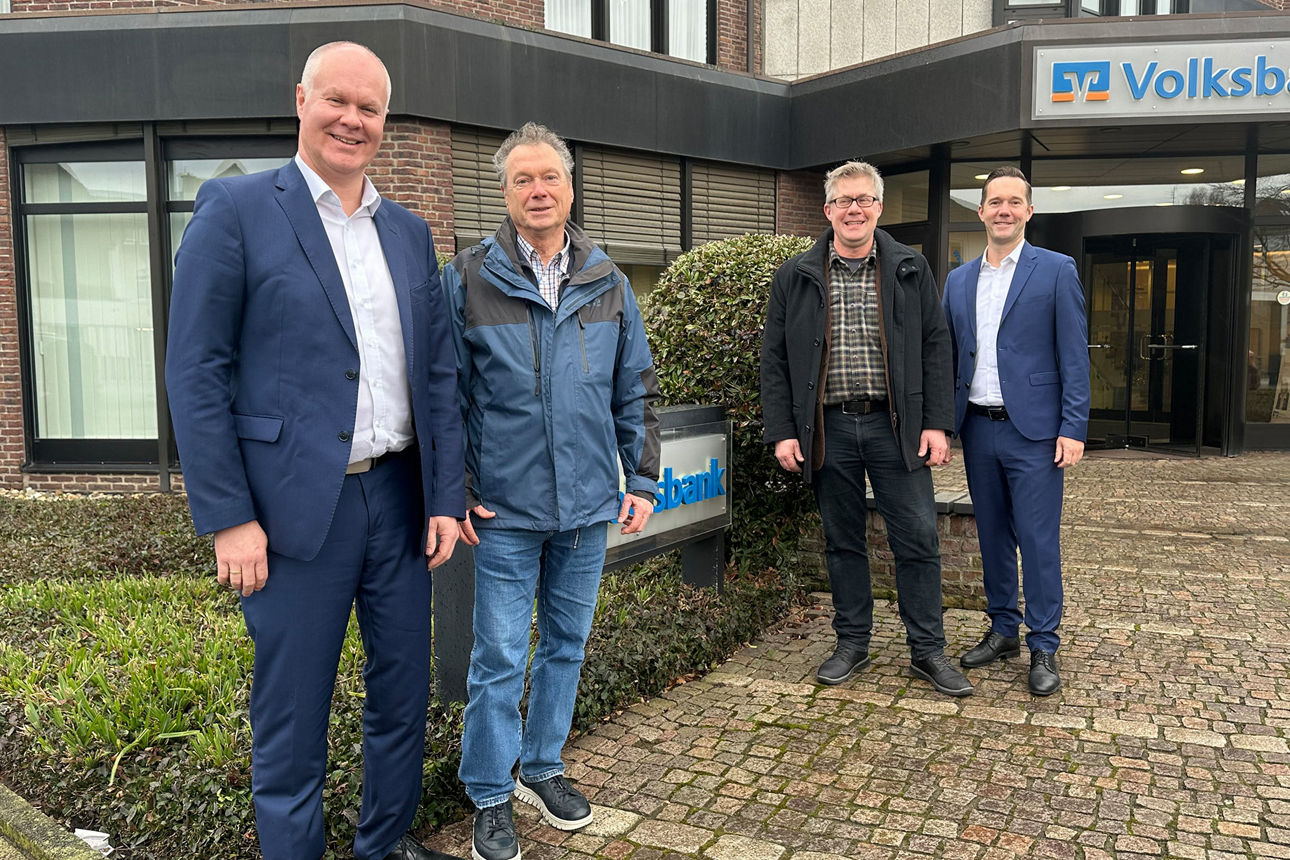 Vier Männer stehen vor einem Backsteingebäude mit einem Schild, auf dem „Volksbank“ steht.