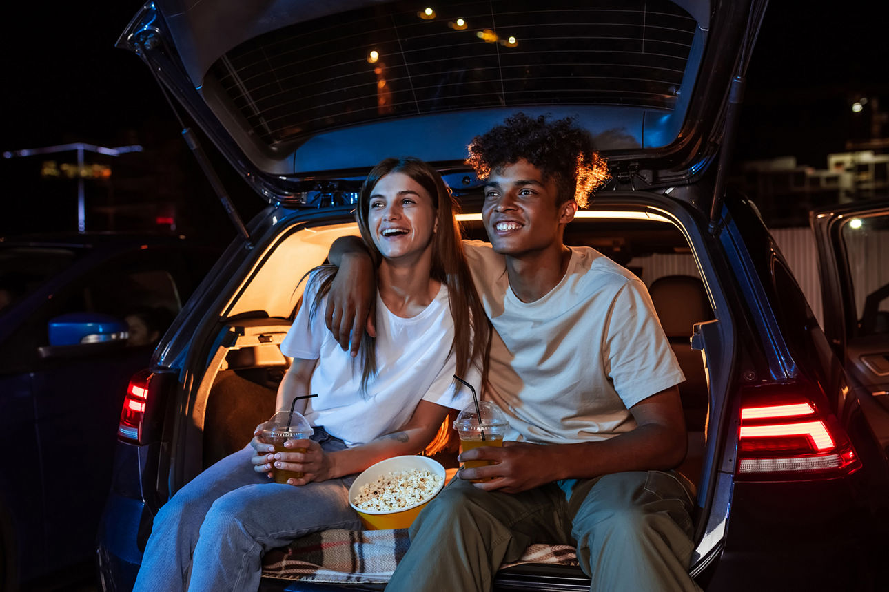 Junges Paar sitzt vor einem Auto und schaut einen Film im Open Air Kino. 