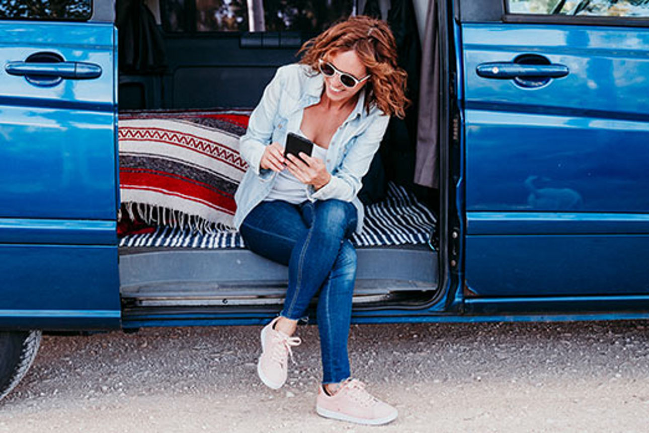 Eine Frau sitzt in der geöffneten Tür eines Vans und schaut auf ihr Smartphone