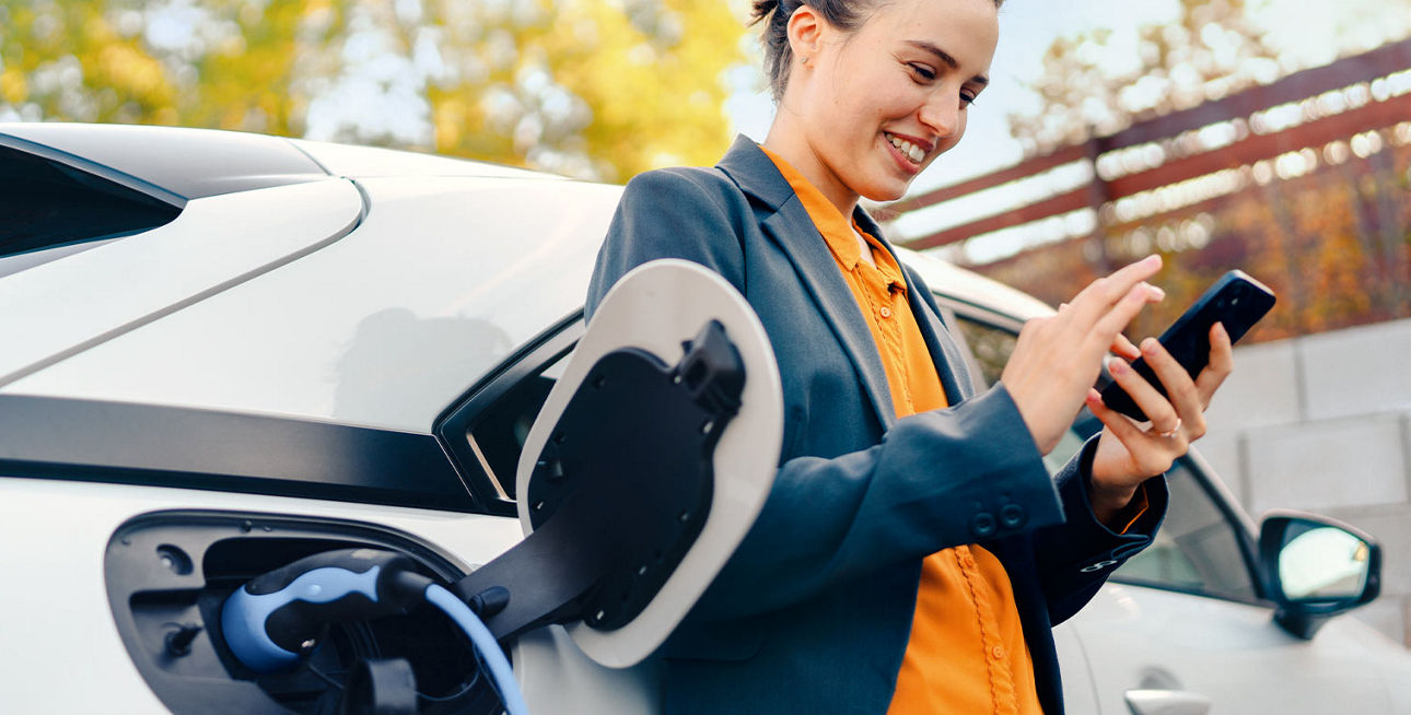 Ein junge Frau steht vor einem E-Auto und bedient ihr Smartphone
