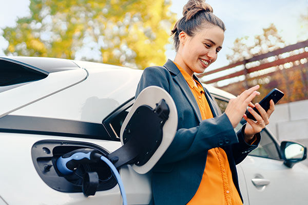 Junge Frau lehnt mit Smartphone an E-Auto