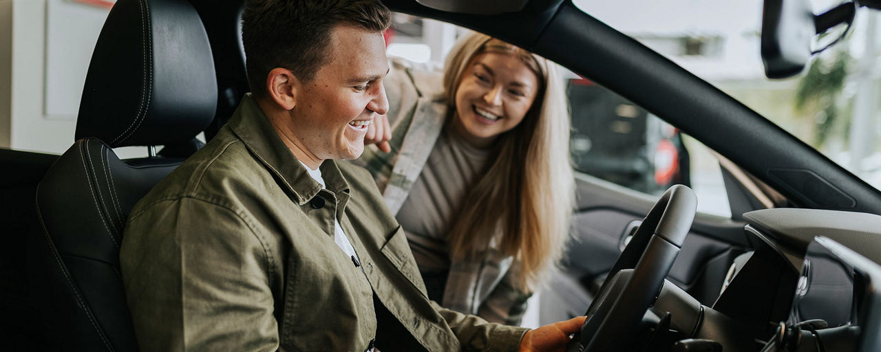 Ein junger Mann sitzt in einem Auto und eine junge Frau steht neben ihm und schaut ihn lächelnd an