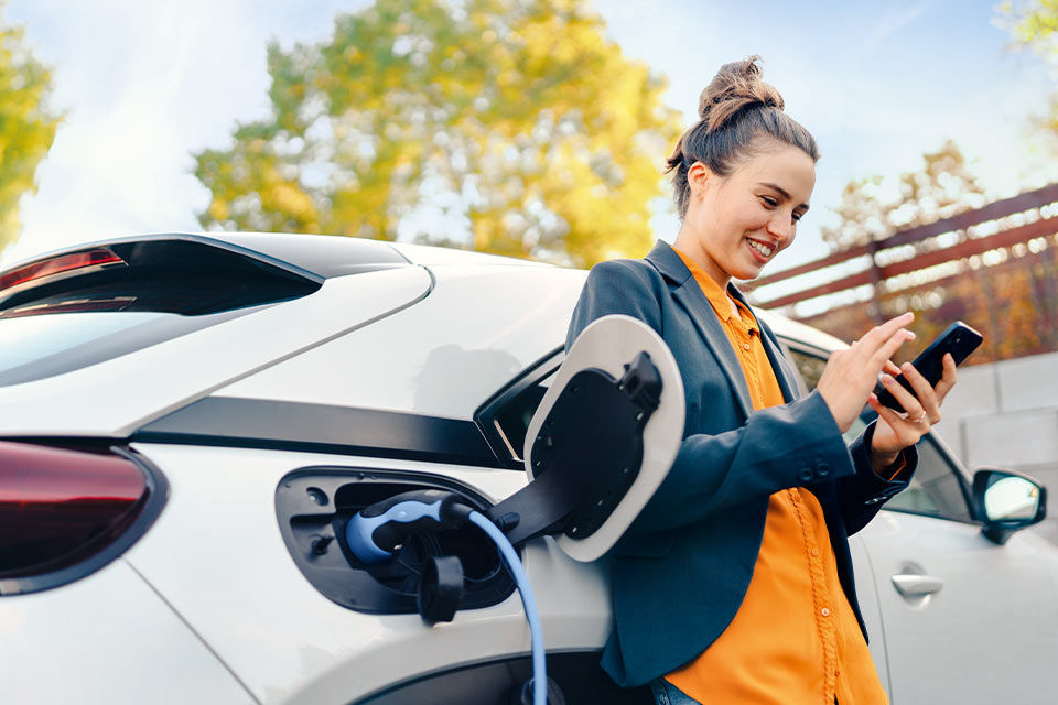 Eine junge Frau steht vor einem E-Auto und bedient ihr Smartphone