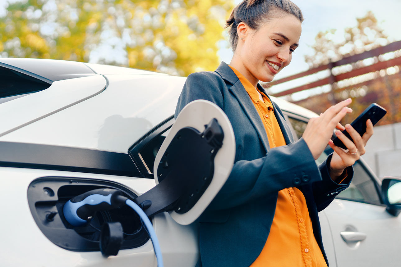 Ein junge Frau steht vor einem E-Auto und bedient ihr Smartphone