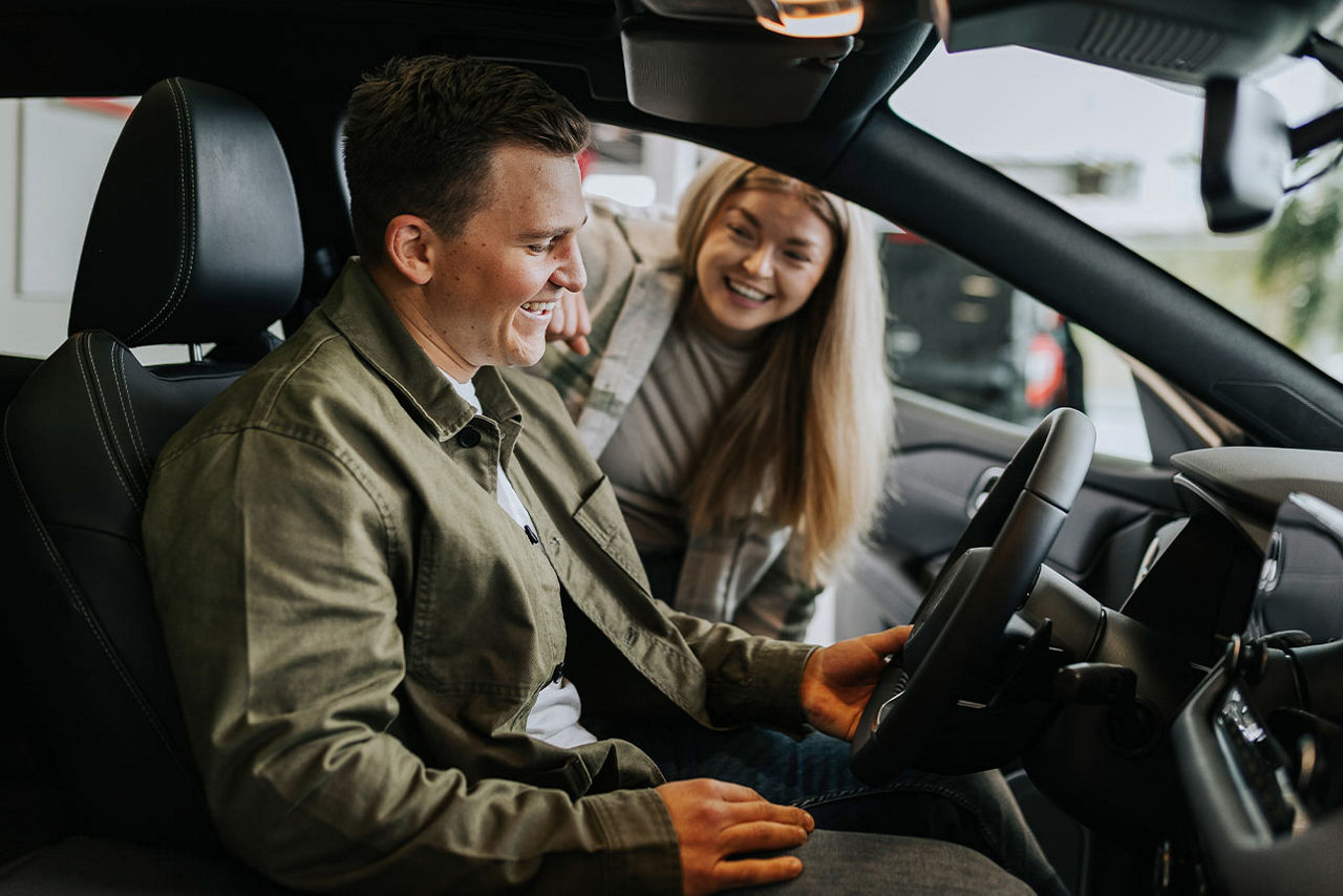 Ein junger Mann sitzt in einem Auto und eine junge Frau steht neben ihm und schaut ihn lächelnd an
