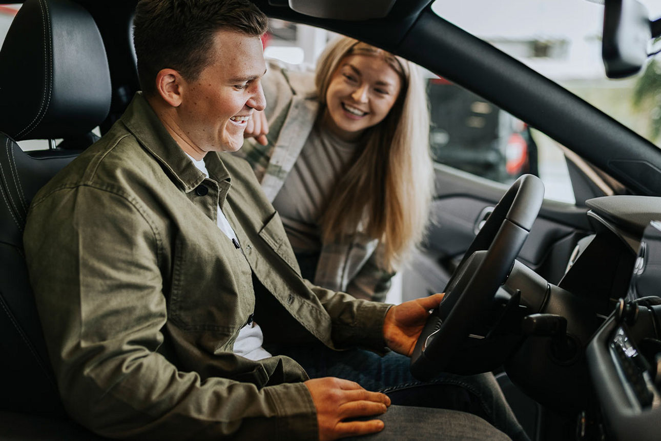 Ein junger Mann sitzt in einem Auto und eine junge Frau steht neben ihm und schaut ihn lächelnd an