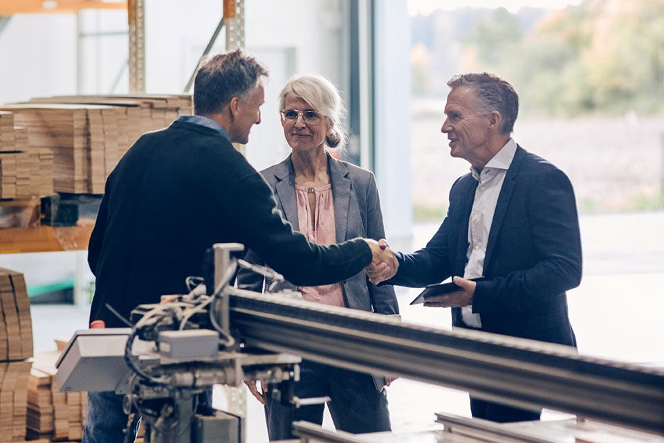 Zwei Männer und eine Frau begrüßen sich mit Handschlag in einer Produktionshalle