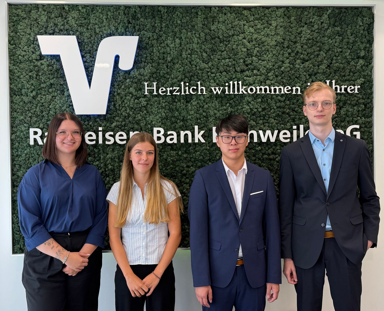 Zwei Frauen und zwei Männer in Geschäftskleidung stehen gemeinsam vor einer begrünten Wand mit Banklogo.