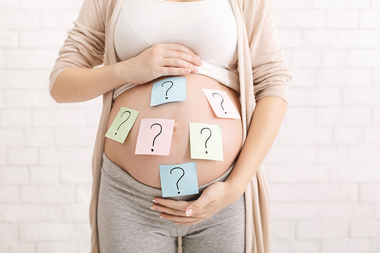 Babynamen Auswahl eines Babynamens: Eine verwirrte schwangere Frau mit Fragezeichen auf Zetteln, die auf ihrem Babybauch kleben.