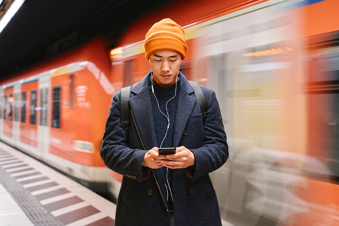 Junger Mann steht am Bahnsteig und schaut auf sein Smartphone