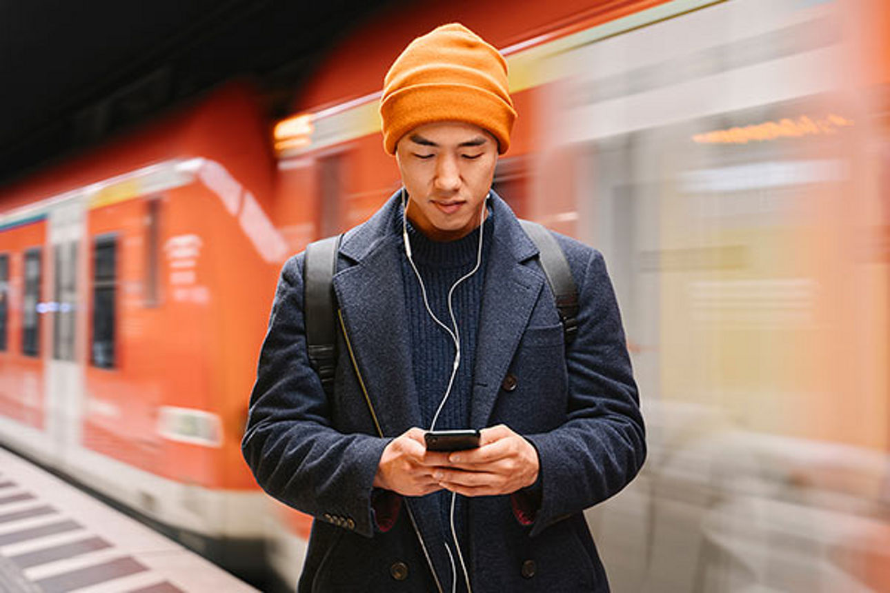 Ein Mann steht mit Smartphone in der Hand und Kopfhörer im Ohr am Bahnsteig