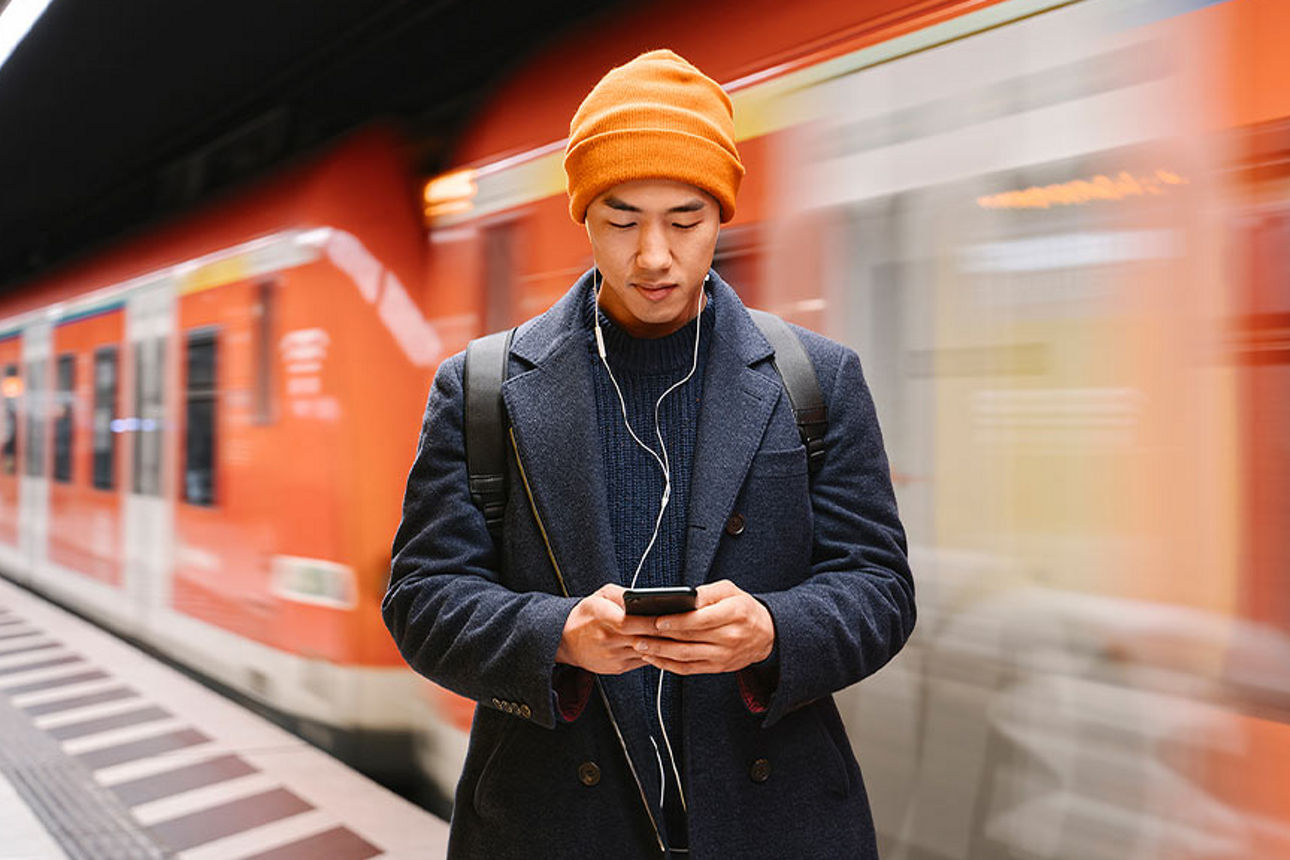 Mann steht in der U-Bahn und schaut auf sein Handy. Hinter ihm rast ein Zug vorbei.