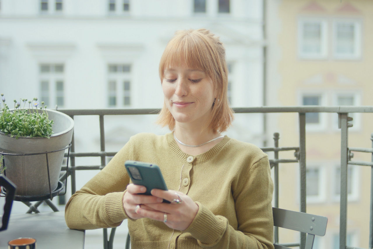 Eine junge Frau sitzt auf dem Balkon und tippt auf ihrem Handy.