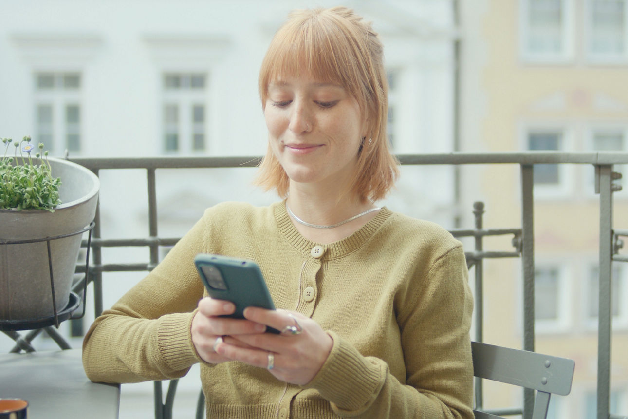 Eine junge Frau sitzt auf dem Balkon und tippt auf ihrem Handy.