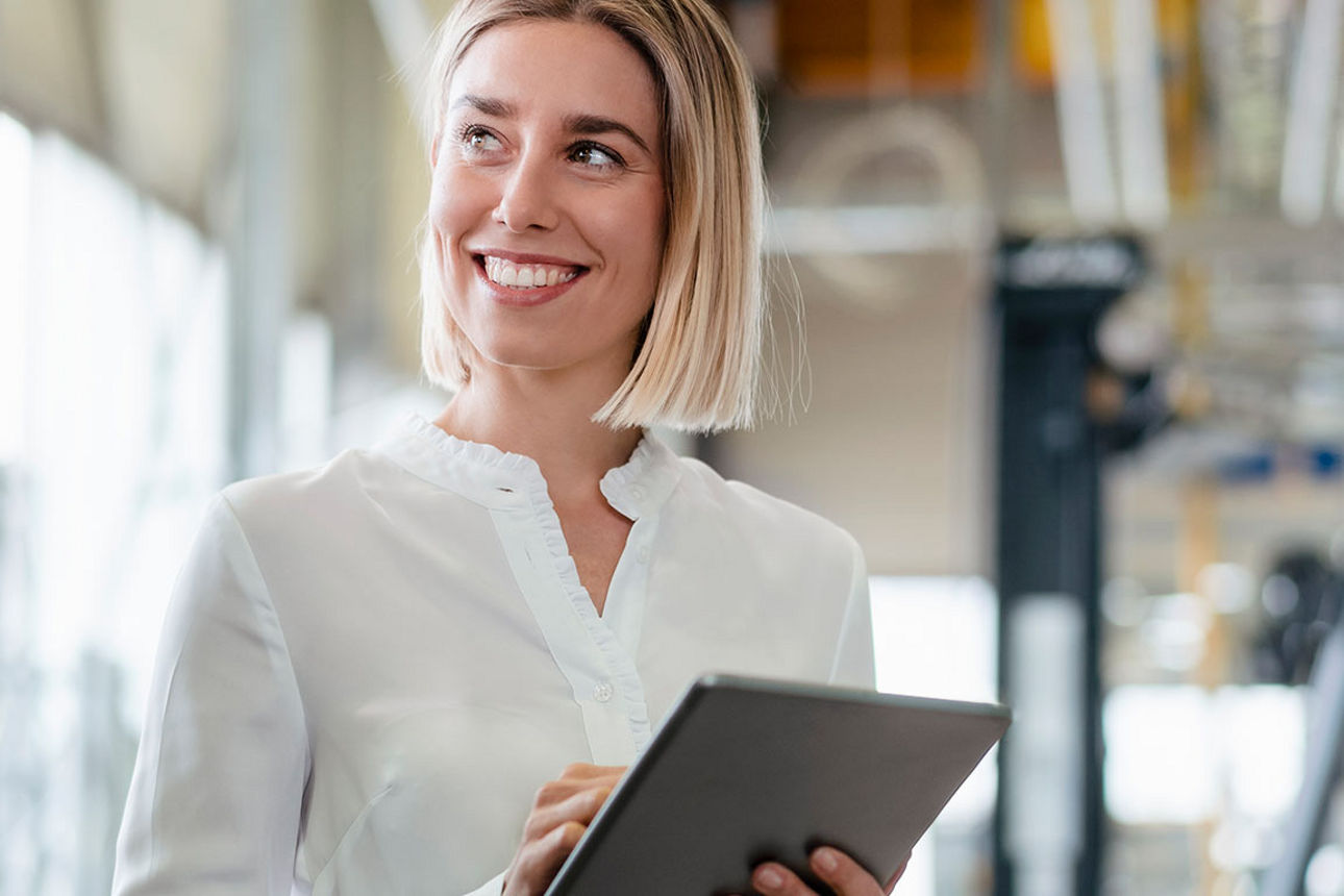  Eine junge blonde Frau mit kurzen Haaren und einem Tablet in der Hand schaut lächelnd zur Seite und notiert auf ihrem Tablet wichtige Erkenntnisse