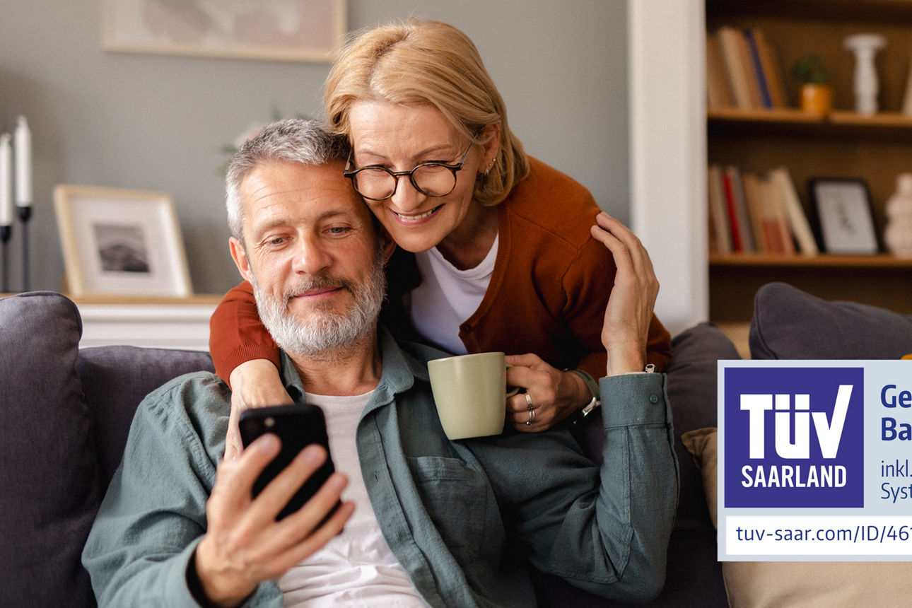 Mann sitzt mit Handy auf dem Sofa. Eine Frau mit Tasse in der Hand steht dahinter und umarmt ihn. Auf dem Bild ist ein TÜV-Siegel "Geprüftes Banking Portal" abgebildet