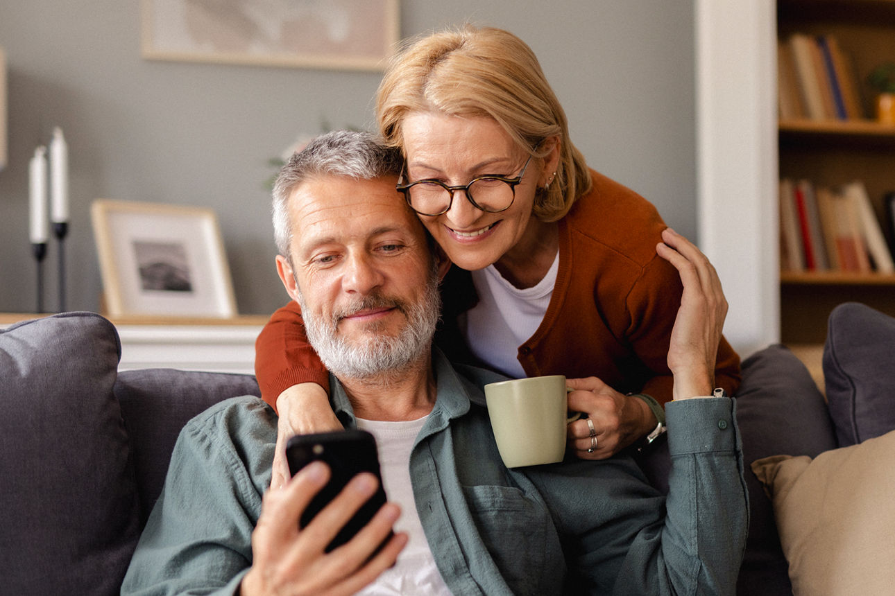 Mann sitzt mit Handy auf dem Sofa. Eine Frau mit Tasse in der Hand steht dahinter und umarmt ihn.