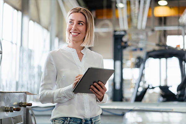Eine junge Frau steht in einer Fertigungshalle und hält lächelnd ein Notebook in der Hand.