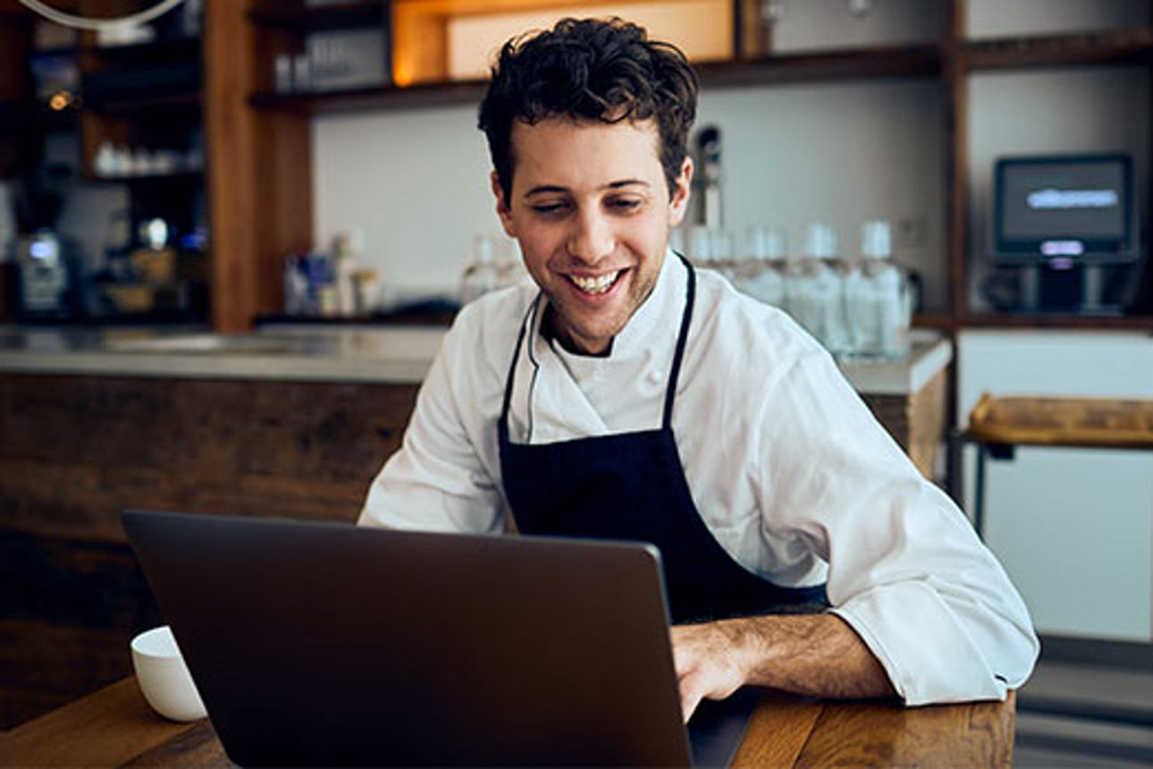 Ein Koch in seinem Restaurant schaut lächelnd auf seinen Laptop