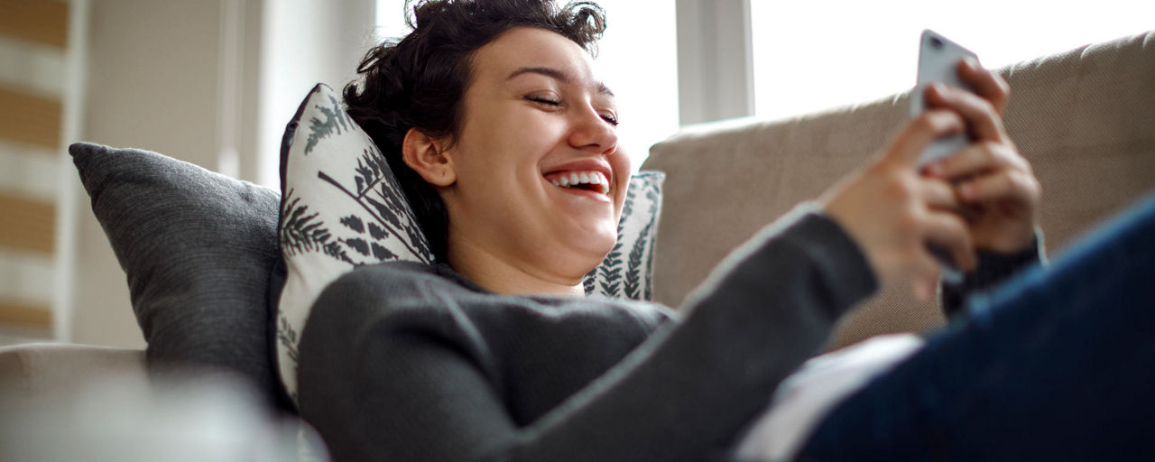 Eine junge Frau liegt auf dem Sofa und schaut lachend auf ihr Handy
