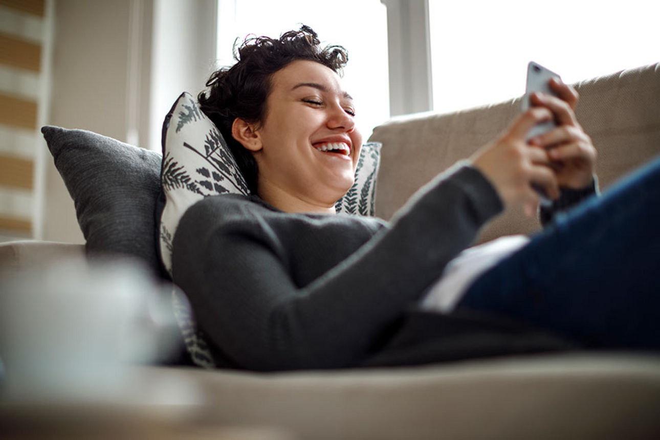 Frau liegt auf einem Sofa und schaut lachend auf ihr Smartphone
