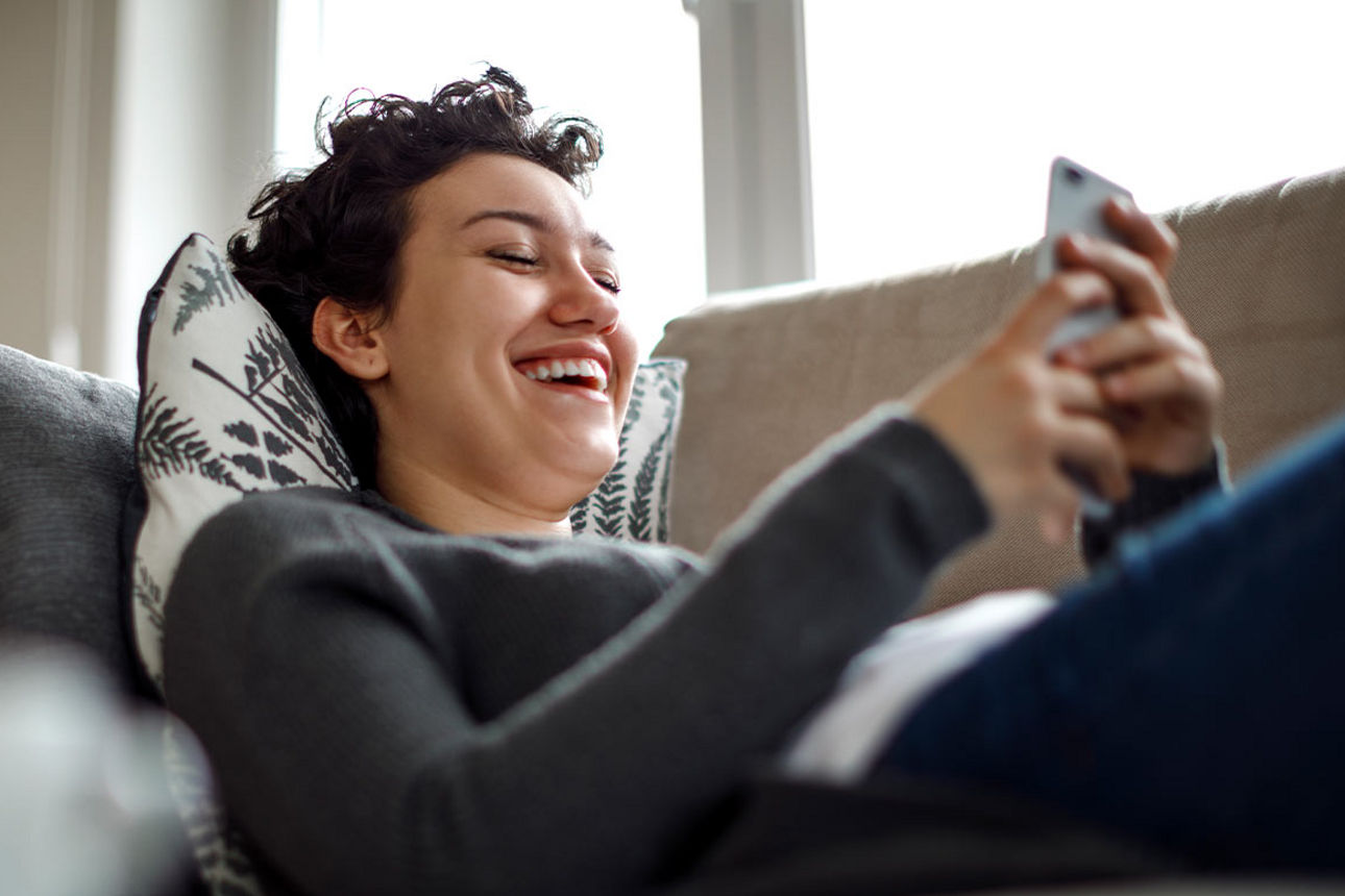 Eine junge Frau liegt auf dem Sofa und schaut lachend auf ihr Handy