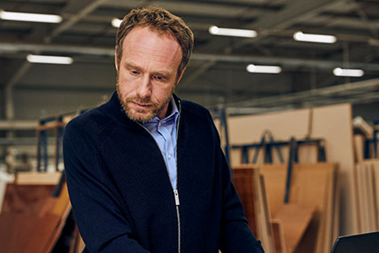 Ein Mann im Pullover steht in einem Lagerhaus und blickt auf einen Laptop