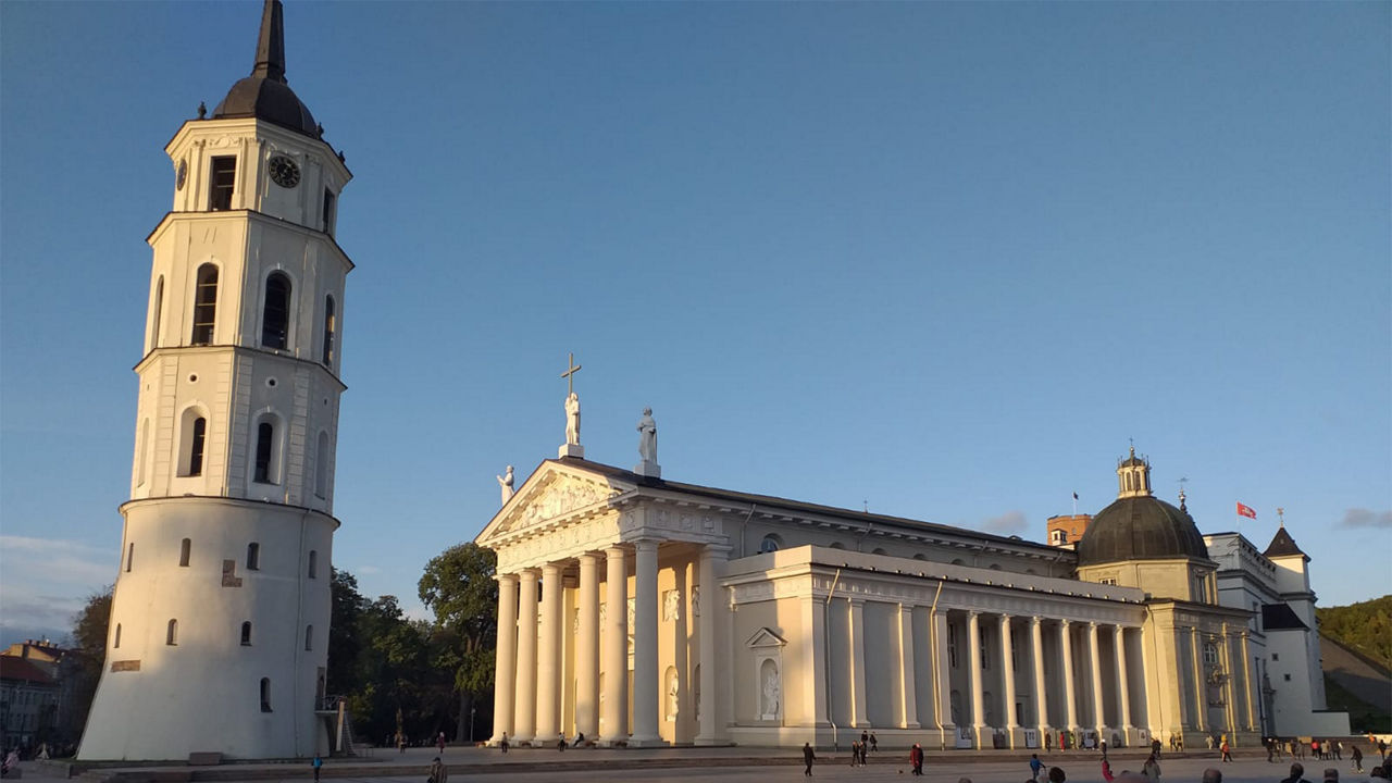 Weißer, freistehender Kirchturm im Sonnenuntergang, neben dem eine Basilika mit Säulen steht