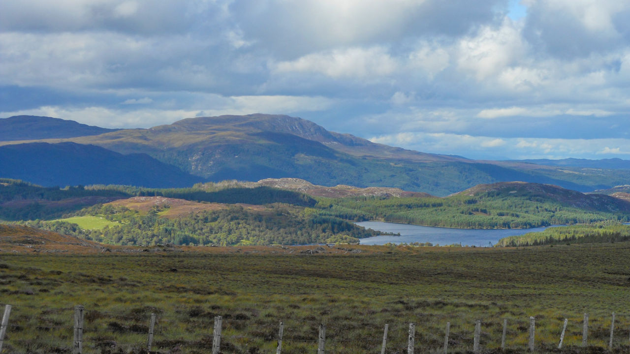 Weidezaun im Vordergrund, hinter dem sich die Highlands mit Wiesen, Wäldern und einem See erstrecken