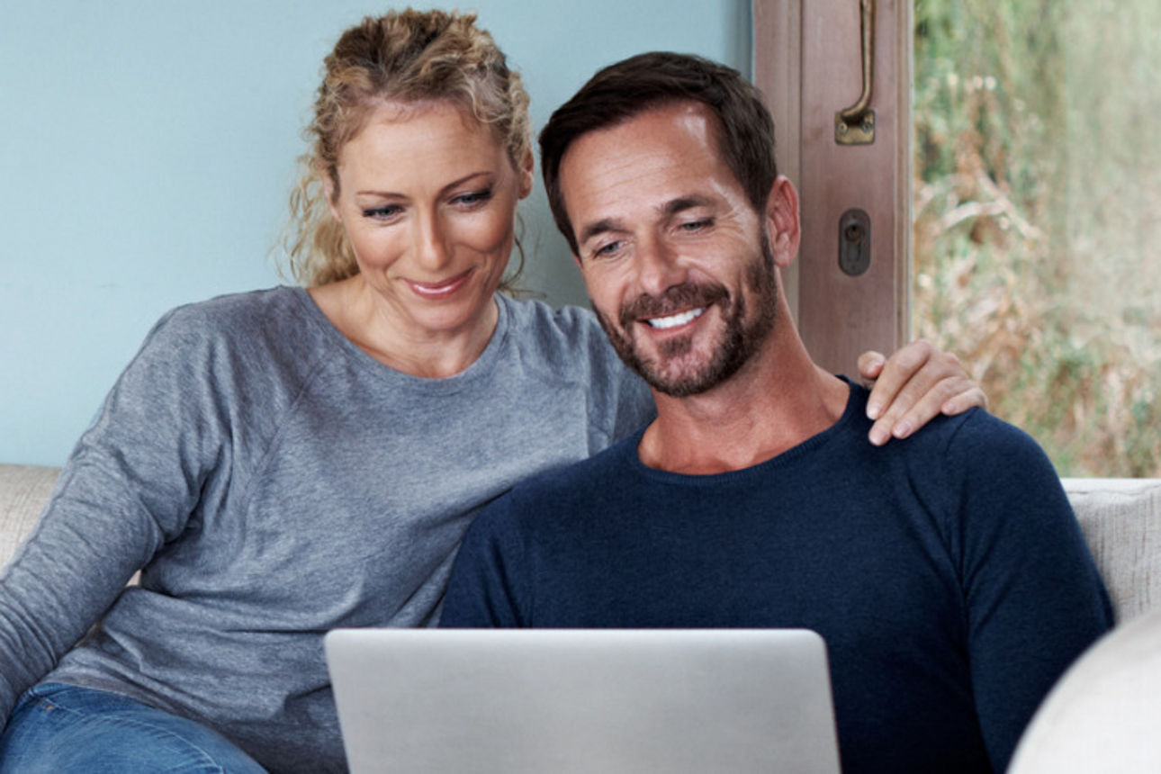 Pärchen sitzt auf der Couch und gucken lächelnd auf Laptop