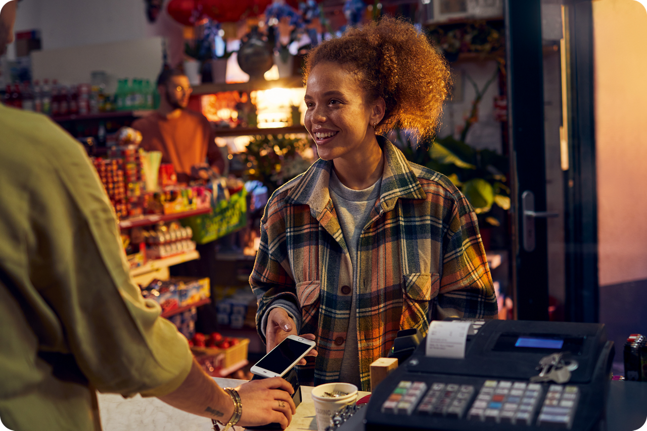 Eine junge Frau hält in einem Kiosk ihr Smartphone hin für eine bargeldlose Zahlung. 