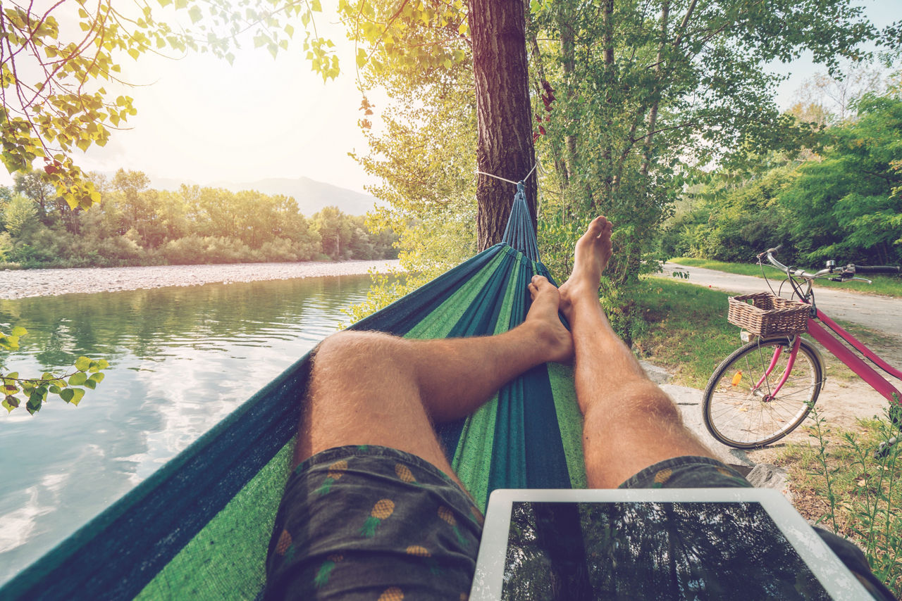 Personal perspective of man lying on hammock by the river using digital tablet in Summer- People travel vacations technology on the go concept